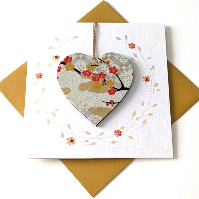 Square handmade greeting card with a gold envelope. The card features a printed delicate floral vine wreath. In the center, a wooden heart ornament is suspended by a metallic gold ribbon. The front of the wooden heart is hand-decoupaged with authentic light-grey Japanese Chiyogami paper featuring red and gold plum blossom patterns.