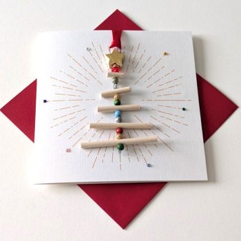 Handmade Christmas card featuring a 3D Christmas tree ornament made of light wooden dowels and colorful beads, topped with a gold star, on white textured paper with a gold sunburst pattern.