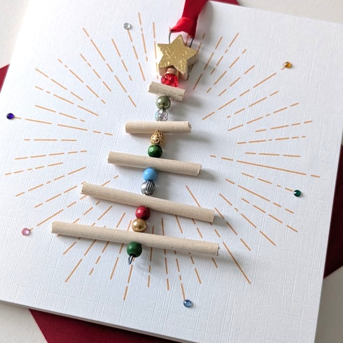 Handmade Christmas card featuring a 3D Christmas tree ornament made of light wooden dowels and colorful beads, topped with a gold star, on white textured paper with a gold sunburst pattern.