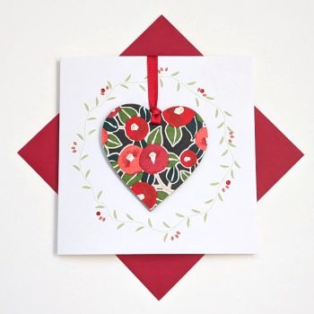 Square handmade greeting card with a red envelope. The card features a printed vine wreath with applied crystals. In the centre, a wooden heart ornament is suspended by a red ribbon. The front of the wooden heart is hand-decoupaged with authentic Japanese Chiyogami paper featuring a red berries and leaf pattern with metallic gold accents.