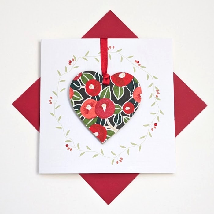 Square handmade greeting card with a red envelope. The card features a printed vine wreath with applied crystals. In the centre, a wooden heart ornament is suspended by a red ribbon. The front of the wooden heart is hand-decoupaged with authentic Japanese Chiyogami paper featuring a red berries and leaf pattern with metallic gold accents.