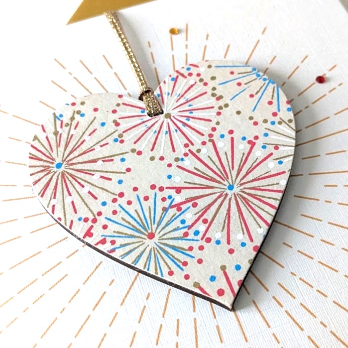 Square handmade greeting card with a gold envelope. The card features a printed starburst effect with applied crystals. In the centre, a wooden heart ornament is suspended by a gold ribbon. The front of the wooden heart is hand-decoupaged with authentic Japanese Chiyogami paper featuring metallic gold, red, blue and white abstract star burst patterns.