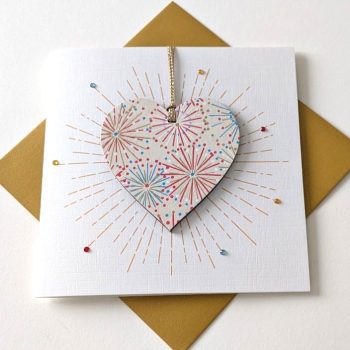 Square handmade greeting card with a gold envelope. The card features a printed starburst effect with applied crystals. In the centre, a wooden heart ornament is suspended by a gold ribbon. The front of the wooden heart is hand-decoupaged with authentic Japanese Chiyogami paper featuring metallic gold, red, blue and white abstract star burst patterns.