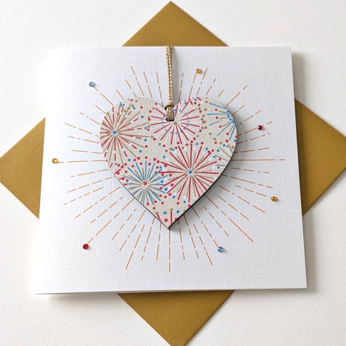 Square handmade greeting card with a gold envelope. The card features a printed starburst effect with applied crystals. In the centre, a wooden heart ornament is suspended by a gold ribbon. The front of the wooden heart is hand-decoupaged with authentic Japanese Chiyogami paper featuring metallic gold, red, blue and white abstract star burst patterns.
