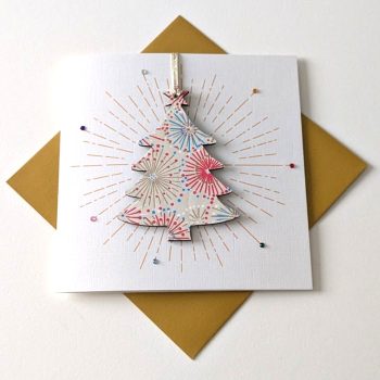 Square handmade greeting card with a gold envelope. The card features a printed starburst effect with applied crystals. In the centre, a wooden tree shaped ornament is suspended by a gold ribbon. The front of the wooden heart is hand-decoupaged with authentic Japanese Chiyogami paper featuring metallic gold, red, blue and white abstract star burst patterns.