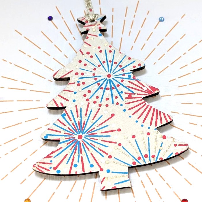 Square handmade greeting card with a gold envelope. The card features a printed starburst effect with applied crystals. In the centre, a wooden tree shaped ornament is suspended by a gold ribbon. The front of the wooden heart is hand-decoupaged with authentic Japanese Chiyogami paper featuring metallic gold, red, blue and white abstract star burst patterns.