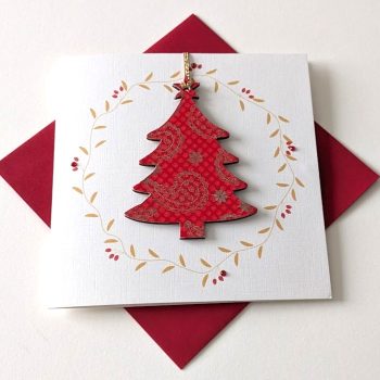Square handmade greeting card with a red envelope. The card features a printed golden leaf wreath with applied crystals. In the centre, a wooden tree shaped ornament is suspended by a red ribbon. The front of the wooden heart is hand-decoupaged with authentic Japanese Chiyogami paper featuring a red dotted print with metallic gold paisley patterns.